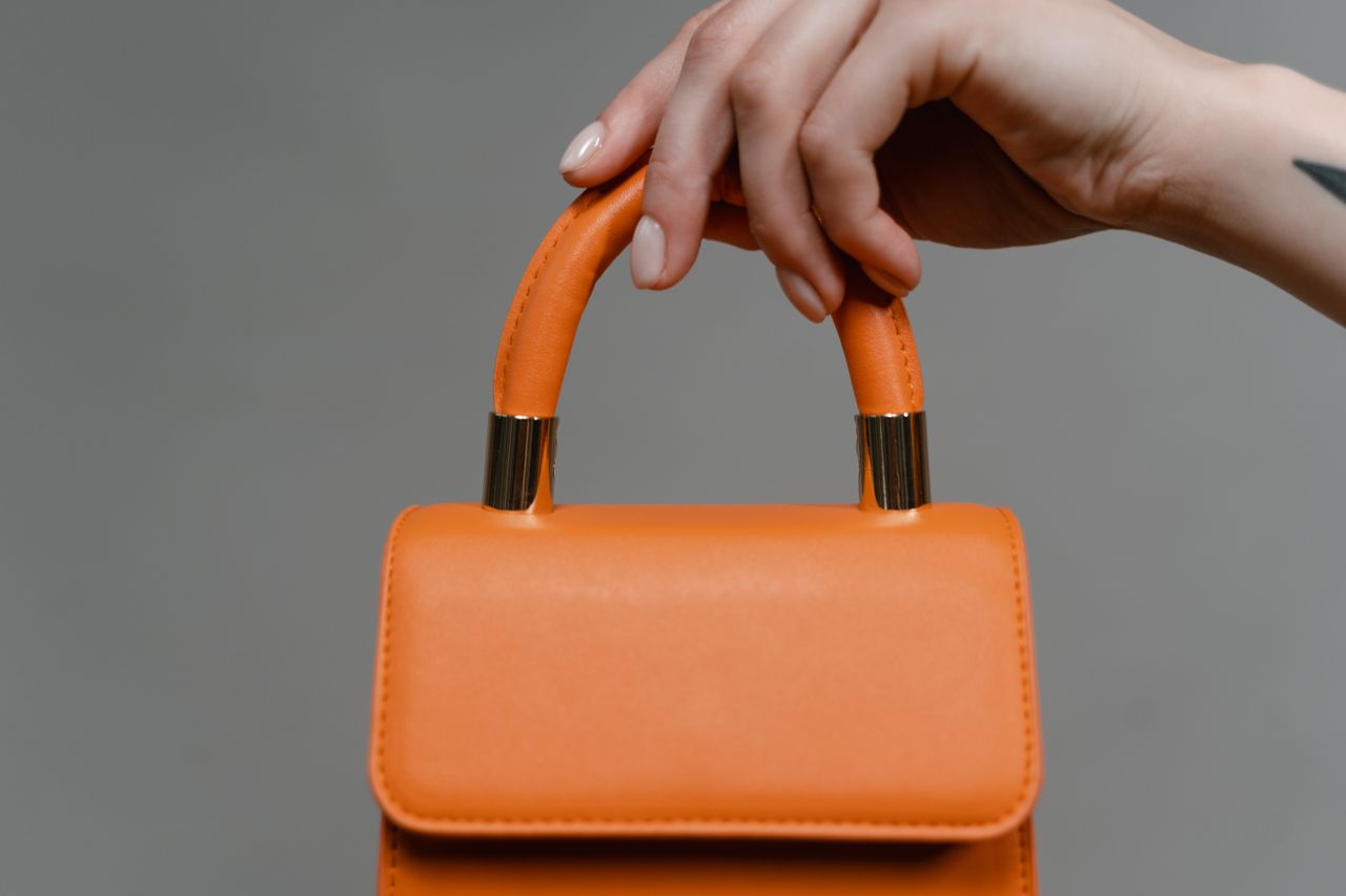 Close-up of a hand holding a stylish orange leather handbag with a gold accent.